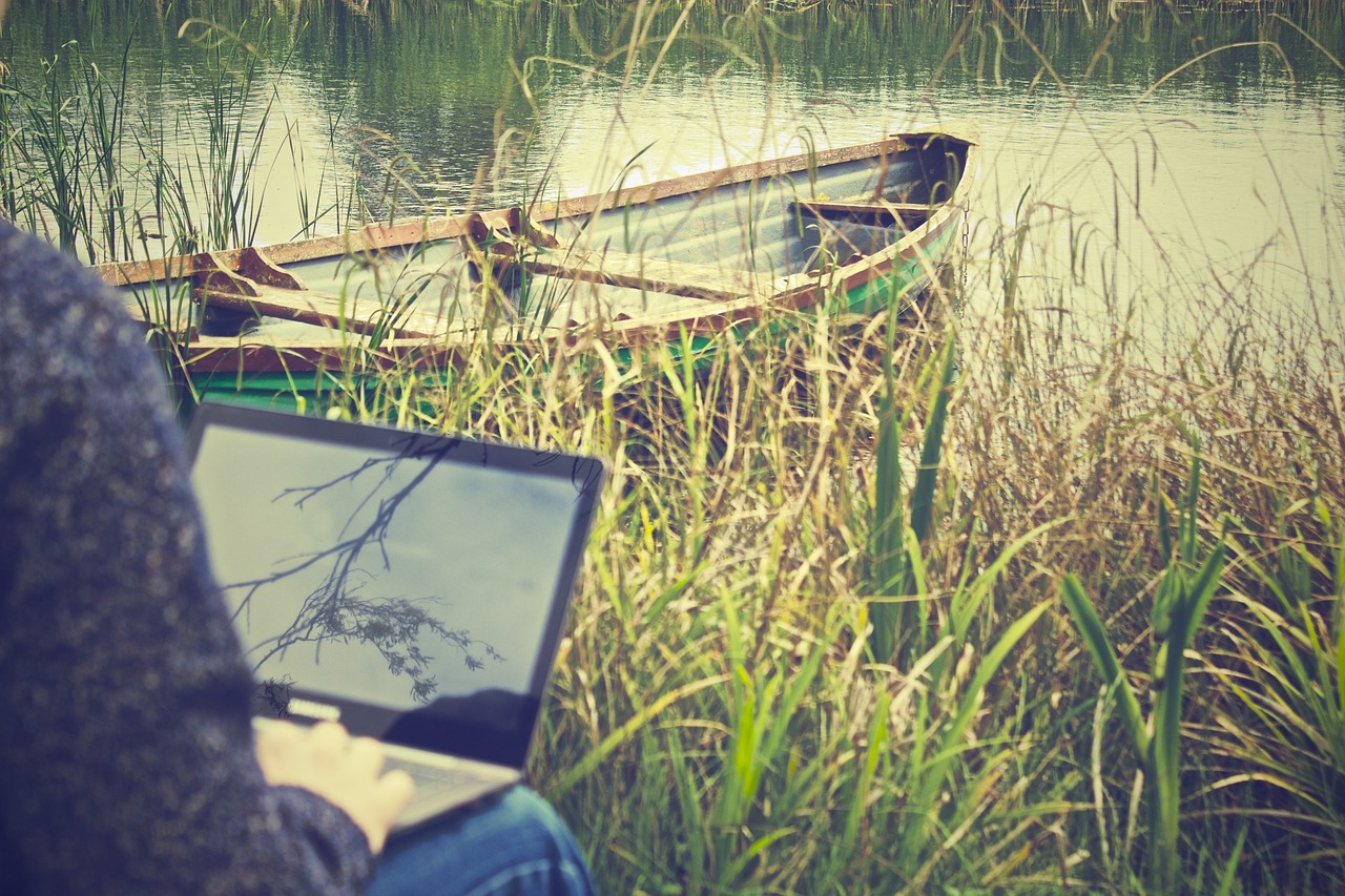 A person sitting outdoors, using a laptop, with a view of a tranquil body of water and a small boat in the background surrounded by tall grass.