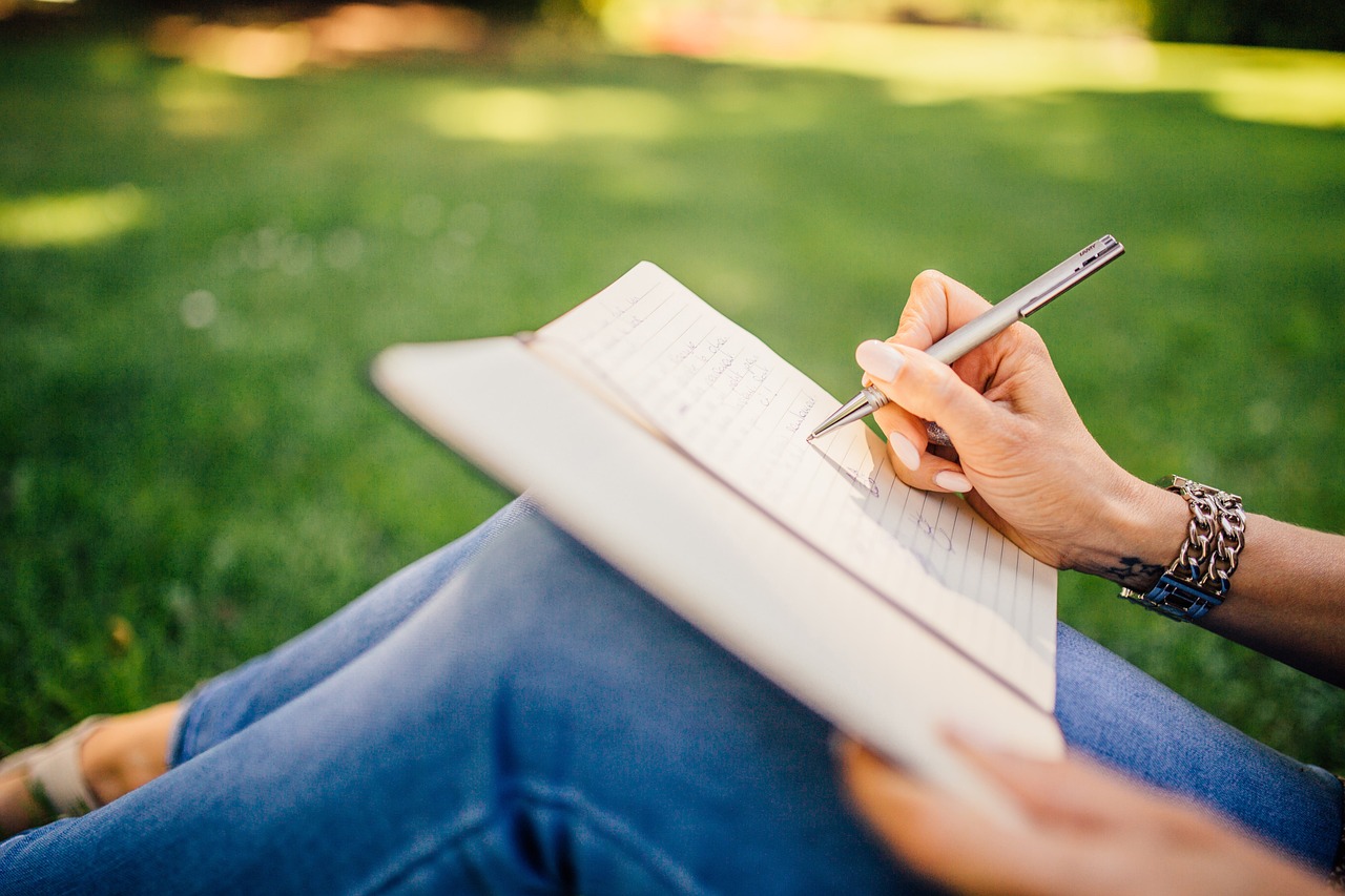 A person writing in a notebook while sitting on grass, holding a pen.