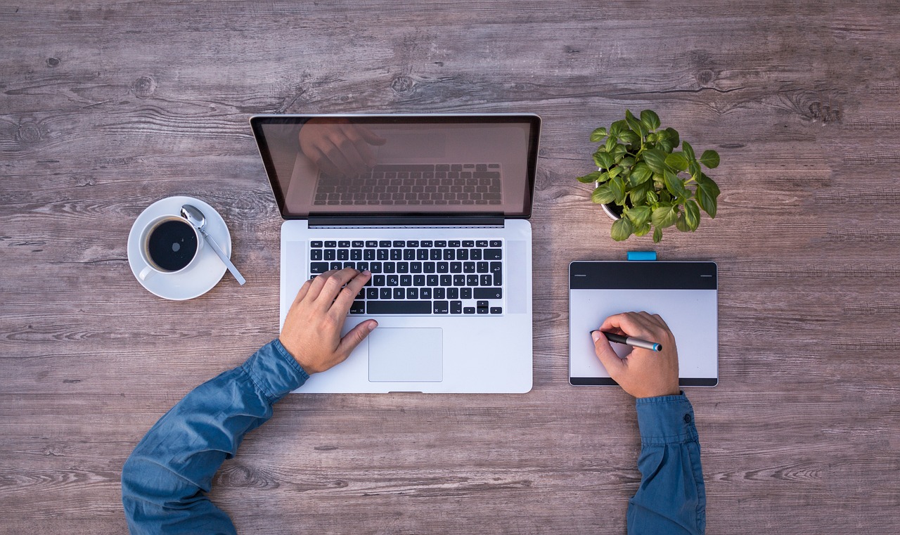 An overhead view of a workspace featuring a laptop, a hand typing on the keyboard, a graphics tablet, and a cup of coffee with a spoon, alongside a small green plant.